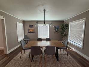 A dining room with a wooden table, four chairs, a chandelier, and two windows with blinds.