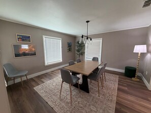 Een eethoek met een houten tafel, zes stoelen, een kamerkleed met patroon en twee ramen met jaloezieën.