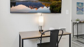 A modern hotel room with a wooden desk, a chair, a mounted painting of a mountain lake, a lamp, and a small table with a plant. - UO•Downtown(.5 mile)•West University•GuestFavorite (Eugene)
