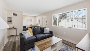 A living room with a dark sofa, a coffee table, a window with blinds, a painting on the wall, and a small table with a lamp. - UO•Downtown(.5 mile)•West University•GuestFavorite (Eugene)