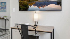 A room with a wooden desk, a chair, a lamp, a painting of a landscape, and a small table with a plant. - UO•Downtown (1/2 mile)•West University•King Suite (Eugene)