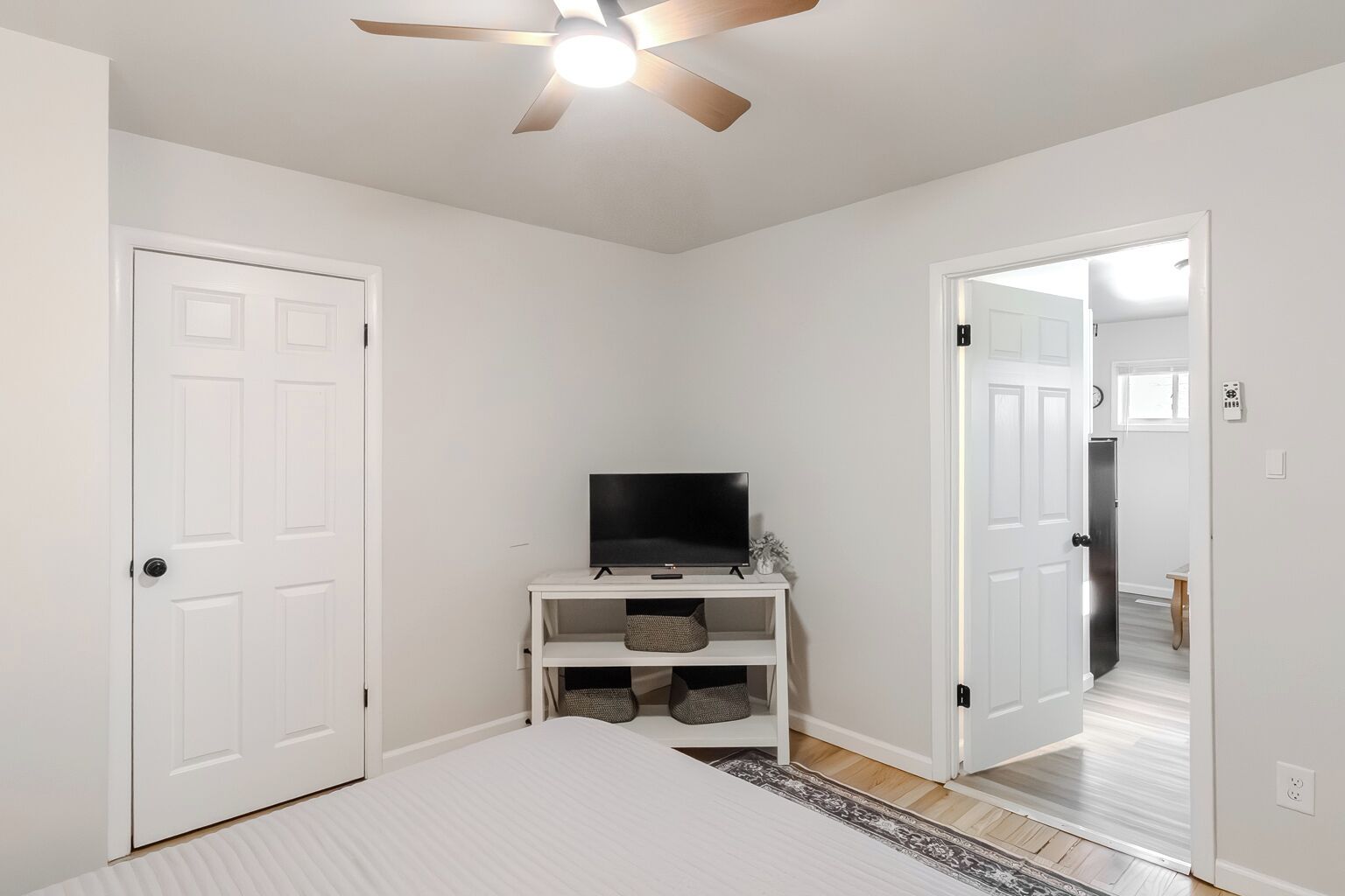 A bedroom with a bed, a TV on a stand, a ceiling fan, and a door leading to another room.