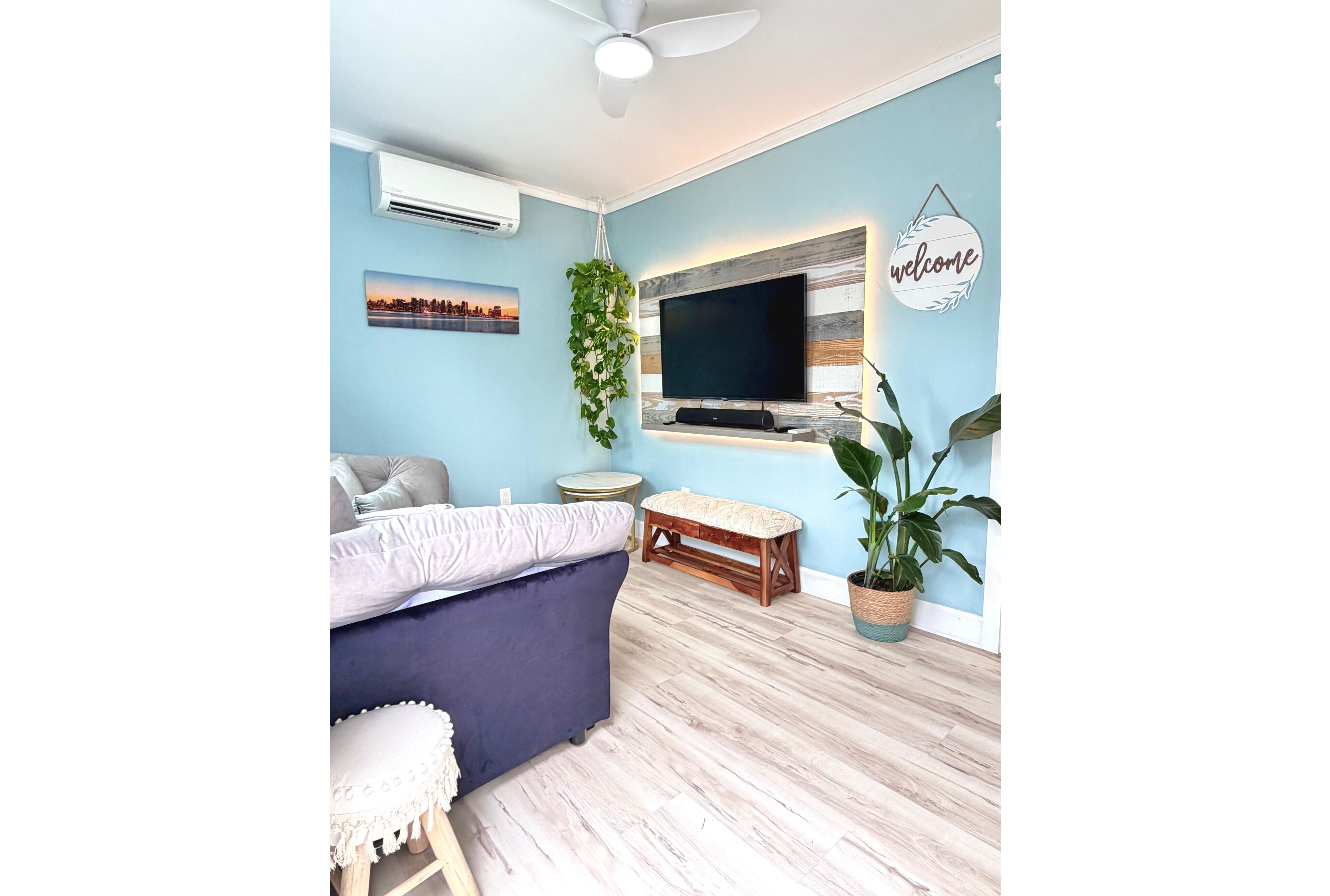 A modern bedroom with a blue wall, a ceiling fan, a flat-screen TV mounted on a wooden panel, a bed with a blue headboard, a small bench, and a potted plant.
