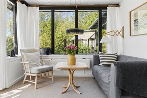 Una habitación acogedora con un sofá gris, una silla de madera, una mesita con un jarrón de flores y un ventanal con cortinas.