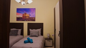 Habitación de hotel con dos camas, una mesita de noche con lámpara, un cuadro de un paisaje urbano y un armario.