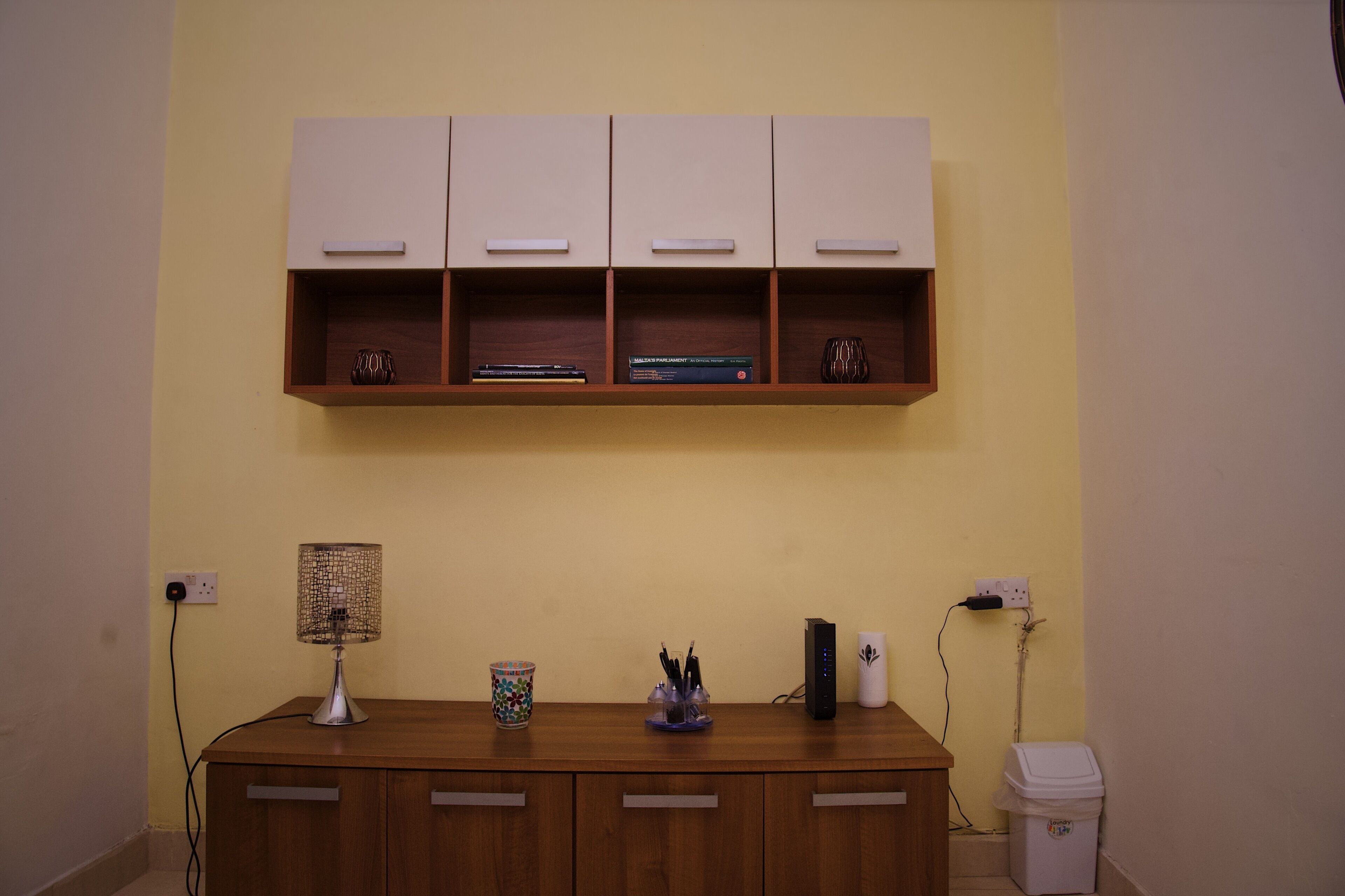 Une pièce avec un bureau en bois, une lampe, une petite étagère remplie de livres et une poubelle.