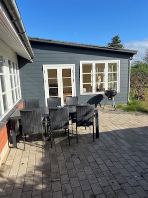 Un patio con una mesa, sillas, una parrilla y una casa con dos ventanas.