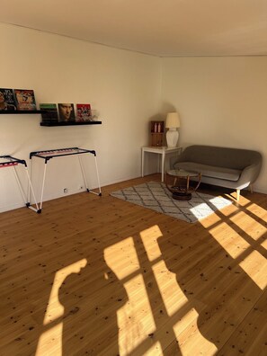 Una habitación con un sofá gris, una pequeña mesa blanca con una lámpara, un estante blanco con libros y objetos decorativos, y un suelo de madera con sombras.