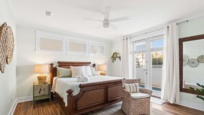Un dormitorio con una cama de madera, una silla de mimbre, un ventilador de techo y vistas a través de un ventanal.