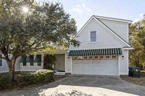 A single-story house with a white exterior, a green awning over the garage, and a tree in the foreground.