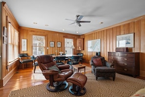A spacious living room with wooden paneled walls, a ceiling fan, a dining area with a table and chairs, and a patterned rug.