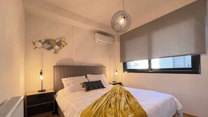 A modern bedroom with a bed, bedside tables, a hanging light fixture, an air conditioning unit, and a window with blinds. - Athenean Modern Escape near Marina Flisvos-E1 (Paleo Faliro)