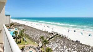 A beach view from a balcony with a pool, beach umbrellas, and a sandy area. - Sandy Key 732 | Spacious Beachfront Condo | Pools, Hot Tub & Tennis | My Beach Getaways (Pensacola)