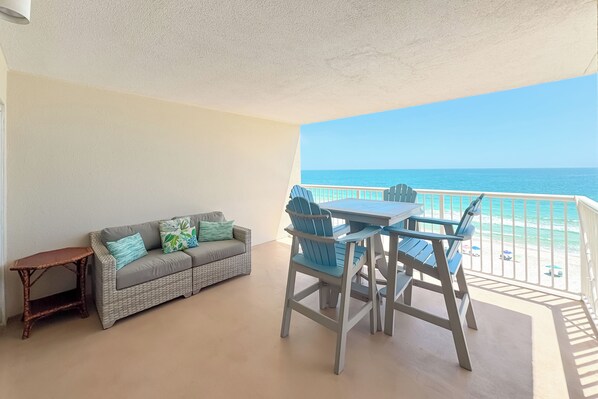 A balcony with a view of the ocean, a table and chairs, a sofa, and a small wooden table. - Sandy Key 732 | Spacious Beachfront Condo | Pools, Hot Tub & Tennis | My Beach Getaways (Pensacola)