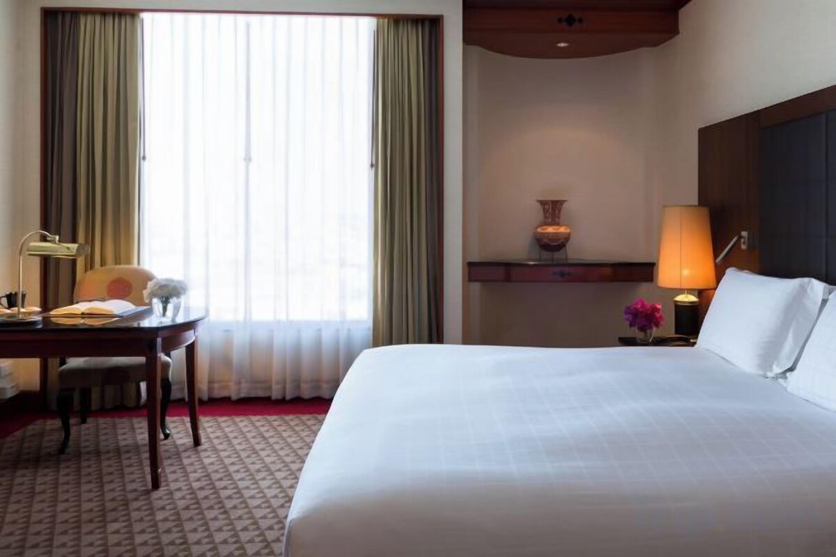 A hotel room with a large bed, a desk with a chair, a lamp, and a vase on a nightstand.