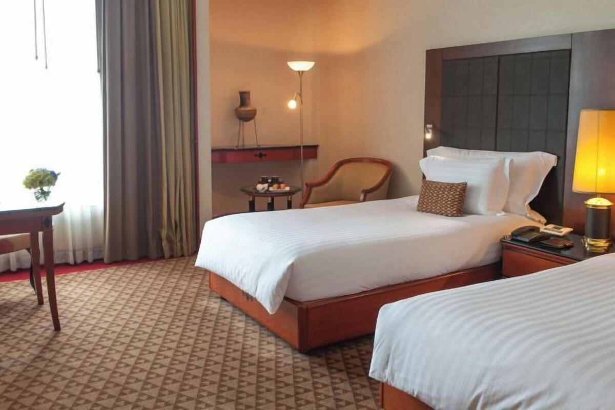 A hotel room with two beds, a desk, a chair, a lamp, and a small table with a vase.
