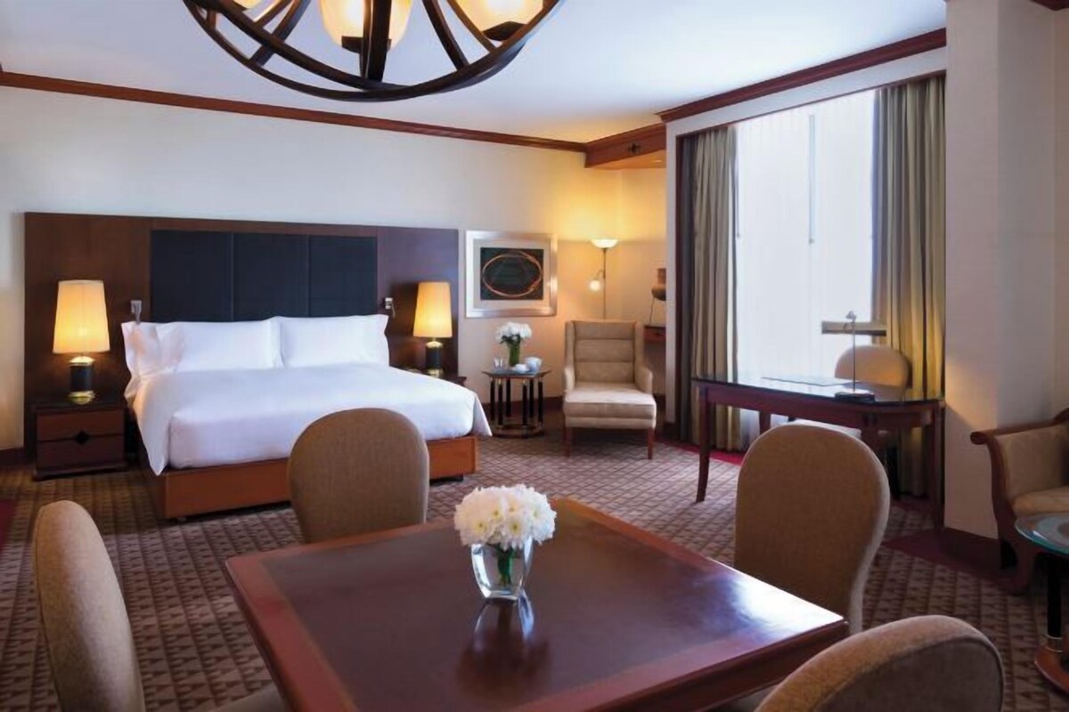 A hotel room with a large bed, two bedside tables with lamps, a desk with a chair, a small table with a vase of flowers, and a large window with curtains.
