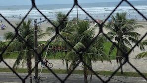 Blick durch ein Maschendrahtgitter auf einen Strand mit Palmen, eine Straße und ein rotes Fahrzeug im Vordergrund.
