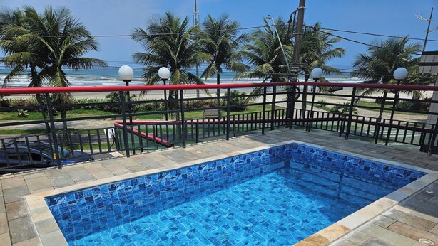 Ein Schwimmbecken mit blau gefliestem Boden, umgeben von einem Metallzaun mit roten Geländern, Palmen und Blick auf den Ozean.