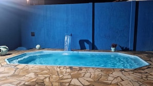 A rectangular swimming pool with a stone-tiled floor, a water fountain, and a blue wall in the background.