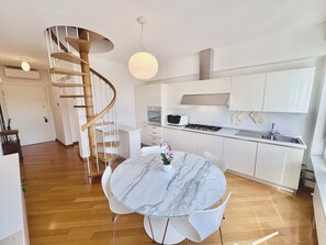 A modern kitchen with white cabinets, a built-in oven, and a gas stove. There is a marble dining table with chairs and a pendant light above it. A spiral staircase leads to another level. - Vittoria Luxury Apartment (Treviso)