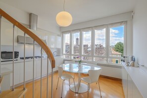 A modern kitchen with a round glass table, white chairs, a wooden staircase, and a view of buildings through large windows. - Vittoria Luxury Apartment (Treviso)