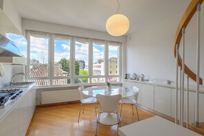 A modern kitchen with a round glass table, white chairs, a gas stove, and a view of buildings through large windows. - Vittoria Luxury Apartment (Treviso)