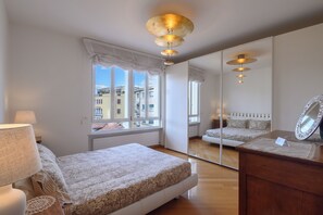 A bedroom with a bed, a bedside table, a mirror, and a window with a view of buildings. - Vittoria Luxury Apartment (Treviso)