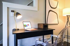 Un bureau avec un ordinateur portable, une lampe de bureau et une chaise transparente, dans une pièce dont le mur est blanc et qui est décorée d’un tableau encadré.