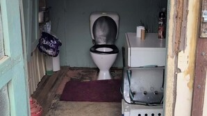 A small, enclosed bathroom with a toilet, a trash bin, and a shelf with toiletries.
