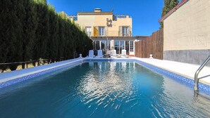 A swimming pool with a tiled edge, surrounded by a wooden fence and a building with large windows in the background. - Cozy home in Guillena (Guillena)