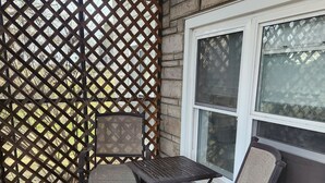 Eine überdachte Terrasse mit Gitterverkleidung, einem Tisch, zwei Stühlen und einem Fenster.