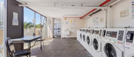 A laundry facility with multiple washing machines, a table with chairs, and a view of the outside through large windows.