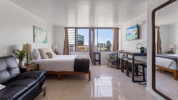A modern hotel room with a bed, a sofa, a dining area, and a large window offering a city view.