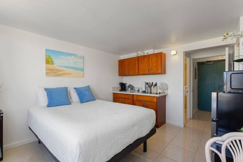 A bedroom with a bed, a painting, wooden cabinets, a kitchen area, and a TV.