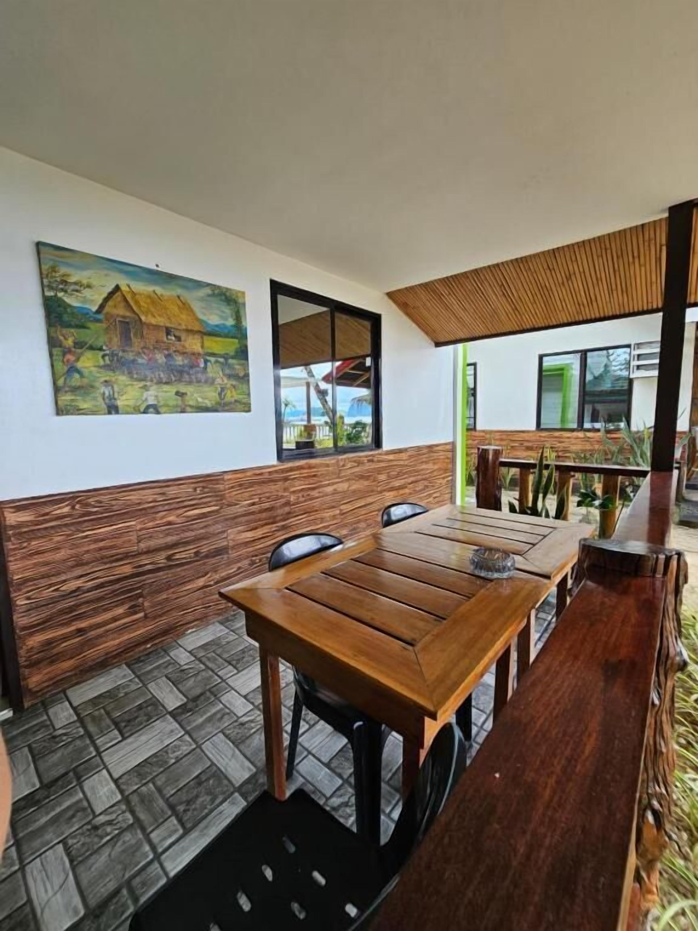 A dining area with a wooden table and chairs, a painting on the wall, and a view of the outside.