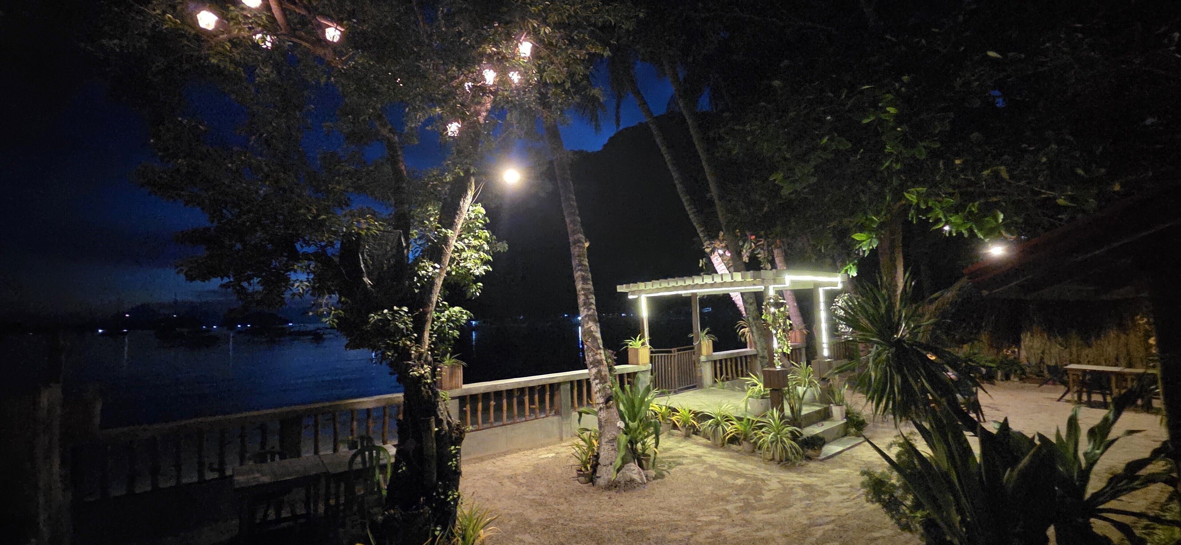 A nighttime view of a waterfront property with a covered seating area, surrounded by trees and plants.