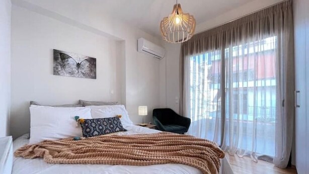 Une chambre à coucher comprenant un lit, une chaise, un luminaire suspendu, un tableau et une fenêtre avec des rideaux.