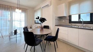 Une cuisine moderne avec des meubles blancs, un coin repas doté d’une table ronde et de chaises, et une grande fenêtre ornée de rideaux légers.