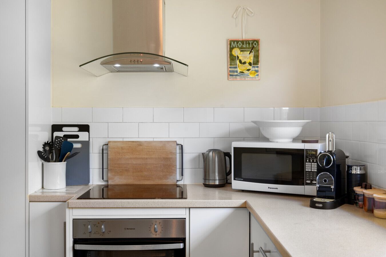 A modern kitchen with white cabinets, a wooden cutting board, a microwave, a kettle, and a coffee machine.