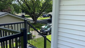 A residential porch with a wooden deck, a black metal railing, and a view of a street with parked cars and a tree.