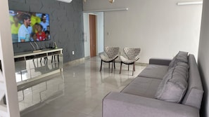 Un salon moderne doté d’un téléviseur à écran plat, d’un canapé gris, de deux fauteuils à motifs et d’un sol carrelé.
