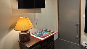 A room with a flat-screen TV mounted on the wall, a wooden nightstand with a lamp and a children's book, and a closed door. - Cozy 2-bedroom condo with AC in pleasant Whitefish (Whitefish)