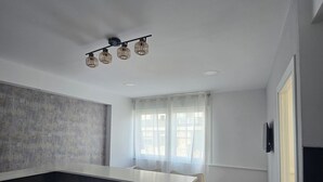 Ein modernes Zimmer mit dunkel gestrichener Akzentwand, einem weißen Schreibtisch, einem Stuhl, einem großen Fenster mit durchsichtigen Vorhängen und einer Deckenleuchte.
