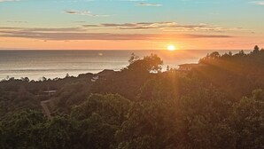 Auringonlasku rannikkomaisemassa, jossa etualalla näkyy talo ja puita.