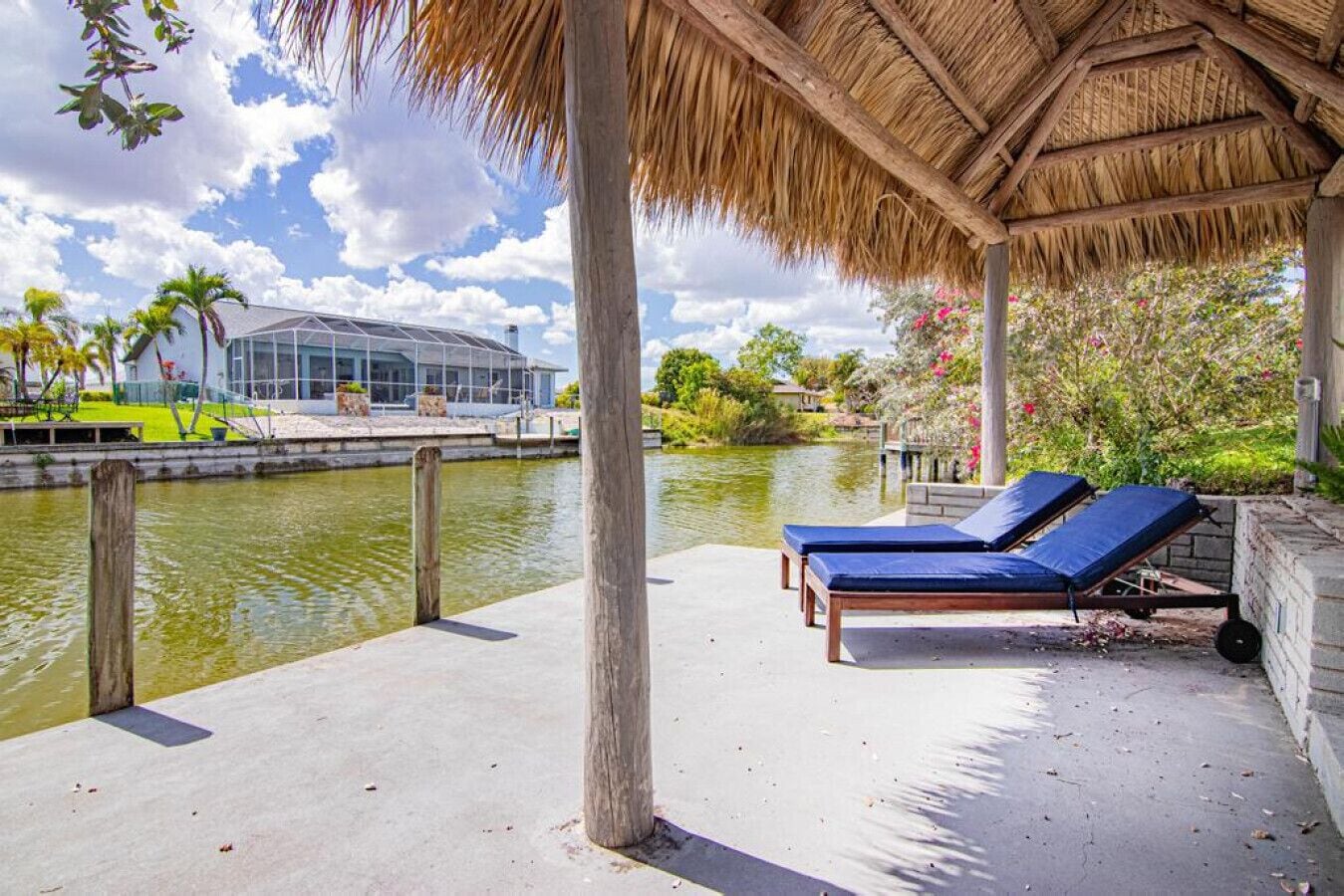 A covered outdoor area with two lounge chairs by a water body, a thatched roof, and a view of a modern building across the water.