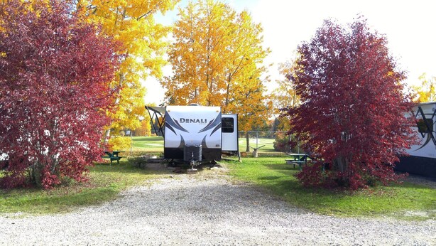 A white RV with 'DENALI' branding parked on a gravel path, surrounded by trees with autumn foliage.