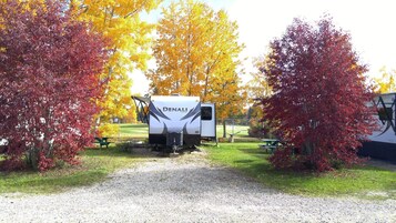 A white RV with 'DENALI' branding parked on a gravel path, surrounded by trees with autumn foliage.