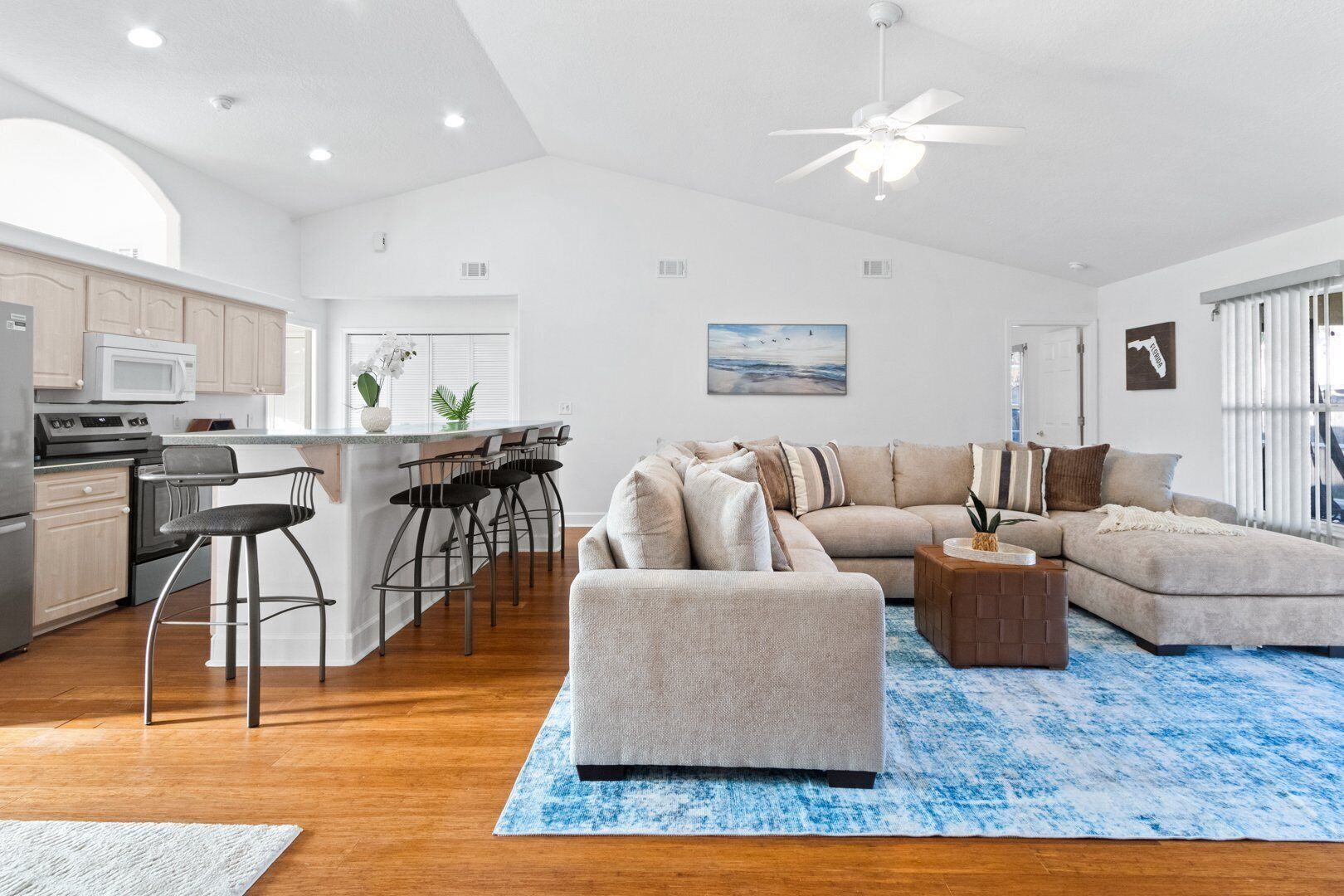 A modern kitchen with a central island, a dining area with bar stools, a living room with a sectional sofa, and a ceiling fan.