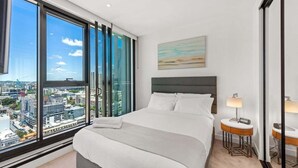 Une chambre d’hôtel moderne avec un grand lit, une vue sur la ville, un tableau au mur et une table de chevet avec une lampe.